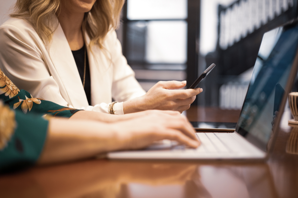 A professional checking her phone with one hand while typing on a laptop, warm office light